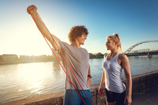 Young fitness couple doing workout with rubber band by the river in a sunset. The man is stretching arms and the woman support him.