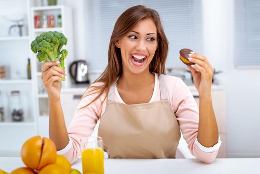 Beautiful young woman in the kitchen decision between broccoli and sweet chocolate cake.