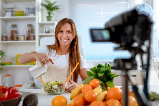 Beautiful young woman filming her blog broadcast about healthy food at  home. She is adding olive oil to vegetable salad.