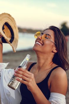 Young woman with a straw hat relaxing at sunset time on the river bank. She is sitting by the river and drinking beer. 