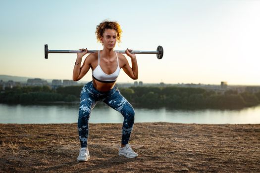Young fitness woman doing workout with barbell by the river in a sunset.