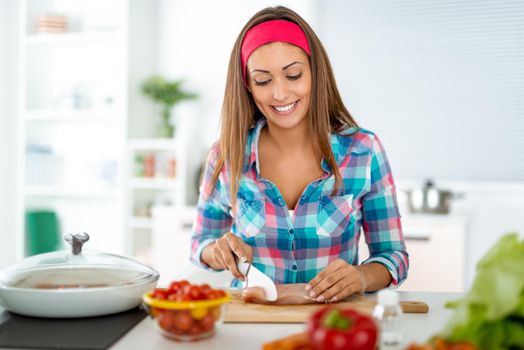 Beautiful young woman making healthy meal in the domestic kitchen. She is cutting chicken meat on the kitchen board.