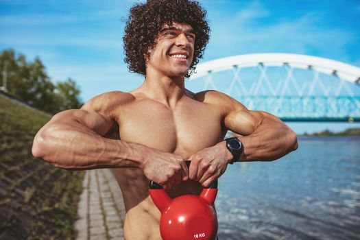 Bodybuilder with six pack, perfect ABS, shoulders, biceps, triceps and chest  is doing strong fit body training with kettlebell, near the river.