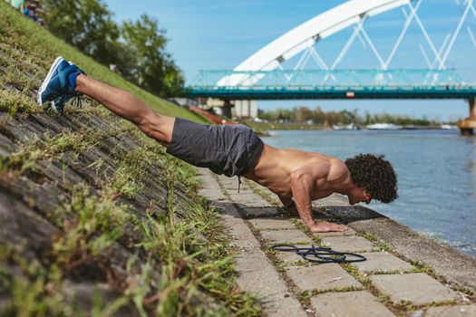 Focused muscular young sport man with naked torso, doing push-ups during outdoor cross training by the river.
