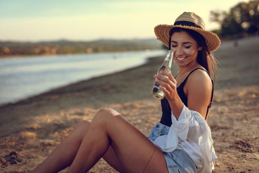 Young woman in straw hat relaxing at sunset time on the river bank. She is sitting on the river send and drinking beer. 