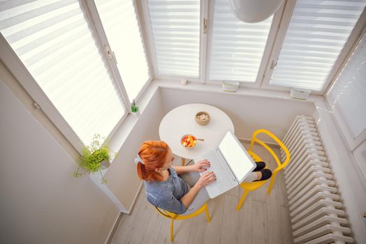 Top view of a beautiful young red haired woman relaxing in the morning, having healthy fruit brekfast and using laptop.