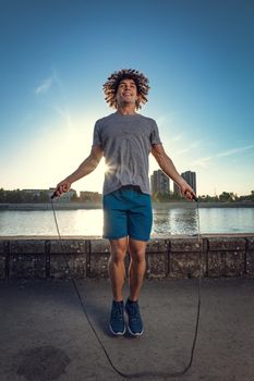 Young handsome sport guy skipping rope near the river against blue sky with sunrise light.