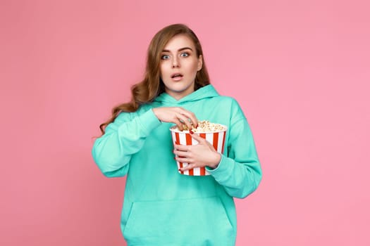 terrified curly brunette girl in sweatshirt watching horror movie , holding bucket of popcorn on pink background