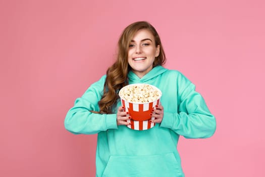 delighted curly brunette woman in sweatshirt holding popcorn isolated on pink background. mockup