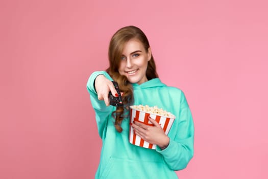 curly brunette woman in turquoise sweatshirt in 3d glasses with remote control holding bucket of popcorn on pink background.