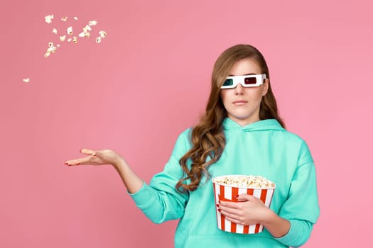 surprised brunette woman in turquoise sweatshirt holding bucket with flying popcorn isolated on pink background