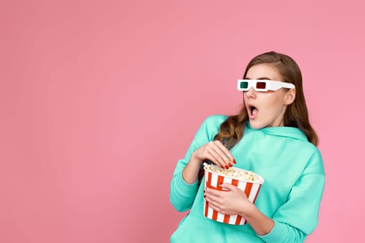 scared terrified curly brunette woman in sweatshirt watching movie film, holding bucket of popcorn on pink background