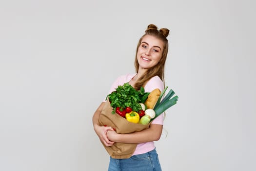 Young smiling caucasian vegetarian woman hold paper bag with vegetables after shopping on gray background