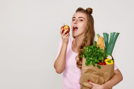 caucasian young woman in pink t-shirt with paper bag with vegetables biting apple. copy space. healthy food for diet