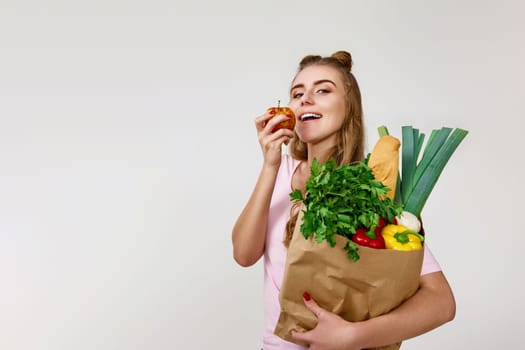 caucasian young woman in pink t-shirt with paper bag with vegetables biting apple. copy space. healthy food for diet
