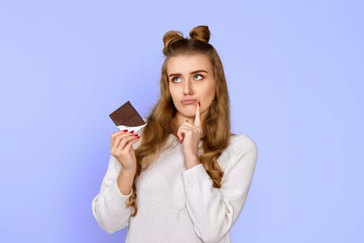 Stylish young woman in sweater holding chocolate bar on violet background