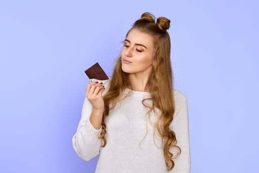 Stylish smiling girl in sweater holding chocolate bar on violet background