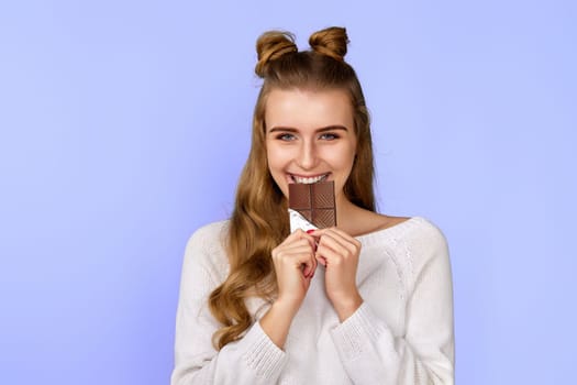 charming happy woman in sweater biting sweet chocolate bar and looking at camera isolated on pastel violet background