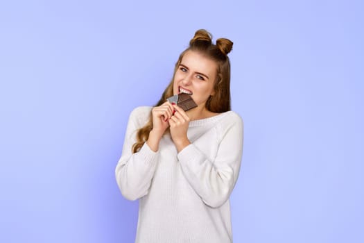 young happy woman in sweater biting sweet chocolate bar and looking at camera isolated on pastel violet background