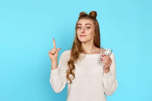 pretty curly brunette woman with drinking glass of water pointing up on blue background