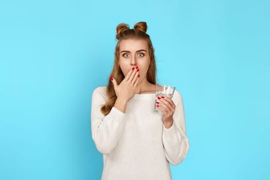 Young surprised curly brunette woman holding glass of water on blue background