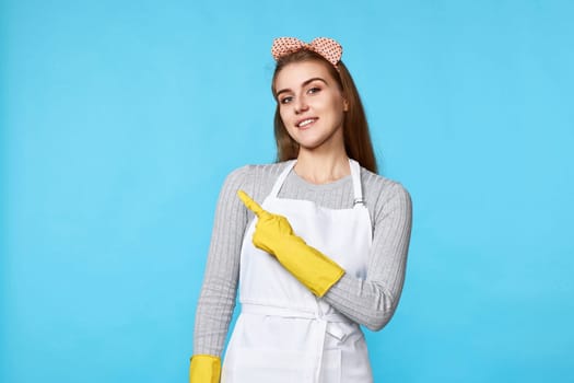 beautiful caucasian woman in yellow rubber gloves pointing fingers aside at copyspace isolated on blue background. cleaning