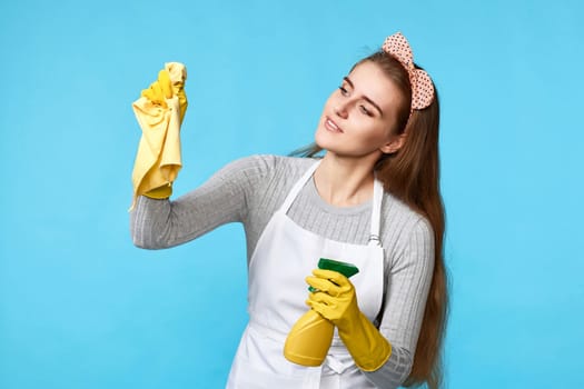 young woman in rubber gloves and cleaner apron cleaning with cleaning rag on blue background.