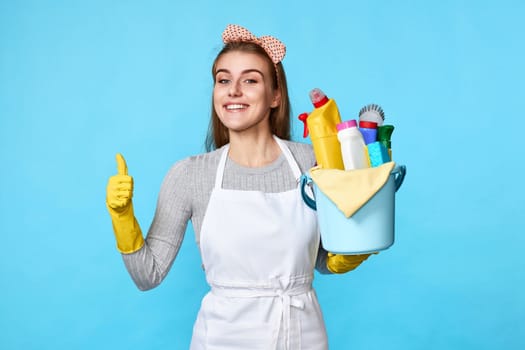 smiling caucasian woman in yellow gloves holding bucket with cleaning supplies showing Ok gesture isolated on blue background.