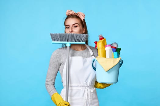 smiling woman in yellow rubber gloves and cleaner apron holding bucket of detergents and broom on blue background.