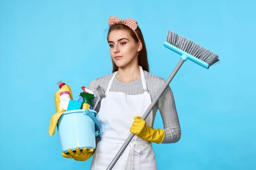 smiling woman in yellow rubber gloves and cleaner apron holding bucket of detergents and broom on blue background.