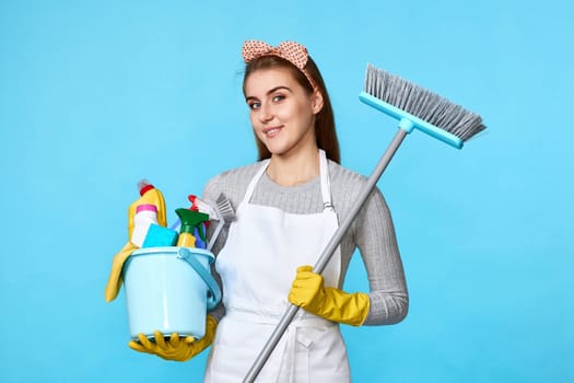 happy woman in yellow rubber gloves and cleaner apron holding bucket of detergents and broom on blue background.