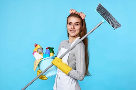 smiling woman in yellow rubber gloves and cleaner apron holding bucket of detergents and broom on blue background.