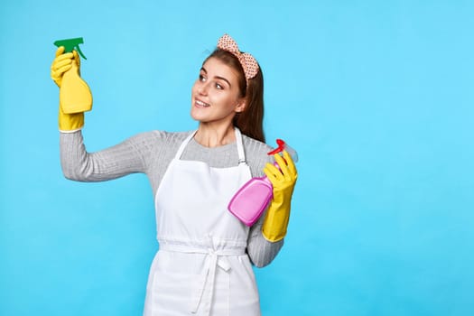 beautiful woman in yellow rubber gloves and cleaner apron chooses between two cleaning products on blue background.