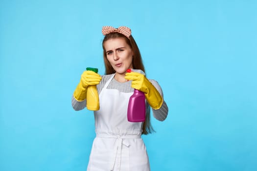 confident cool girl in rubber gloves and cleaner apron cleaning with detergent sprayer on blue background.