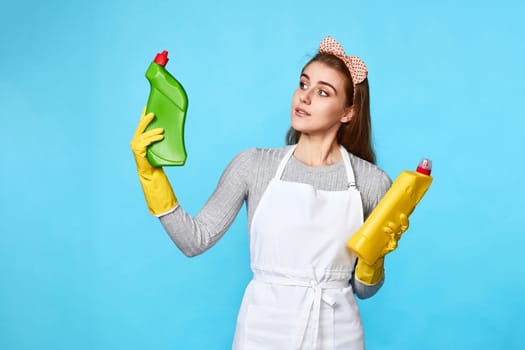 beautiful woman in yellow rubber gloves and cleaner apron chooses between two cleaning products on blue background.