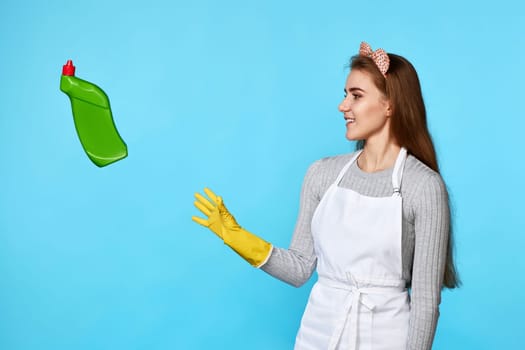 caucasian woman in yellow rubber gloves catches flying bottle of detergent on blue background.