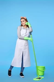 pretty woman in green rubber gloves and cleaner apron washing floor with mop on blue background. Full length