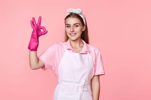 beautiful happy woman in rubber gloves and cleaner apron showing ok sign isolated on pink background. cleaning