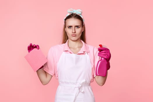 skeptic and nervous, frowning woman in pink gloves and cleaner apron with cleaning rag and detergent sprayer