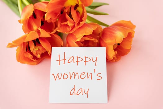 A beautiful bouquet of orange peony tulips along with a white card in the middle with the words happy women's day on a pink background.