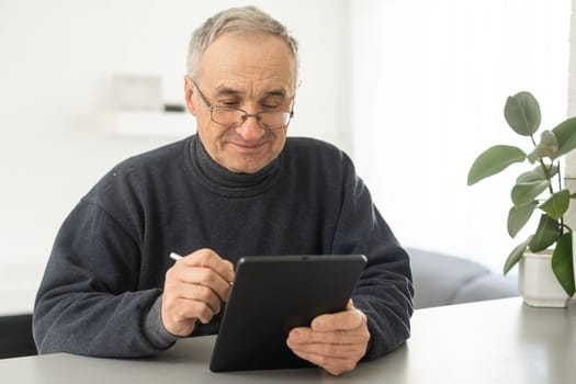 Senior man reading news on digital tablet. Serious mature male using portable computer at home, sitting at table, copy space