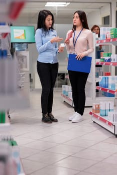 Team of people talking about pills and medicine in pharmacy shop, medical specialist explaining prescription treatment or drugs to client. Asian customer and pharmacist looking at vitamins box.