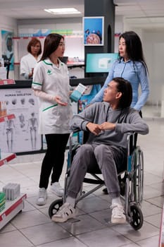 Asian man in wheelchair talking to pharmacist about prescription medicine and pharmaceutical products. Client with disability buying medicinal supplements and vitamins, box of drugs.