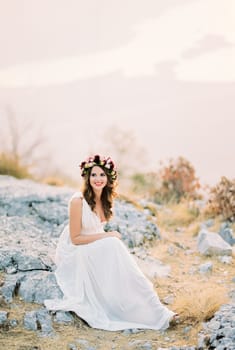 Smiling bride in a flower wreath sits on stones on top of a mountain. High quality photo