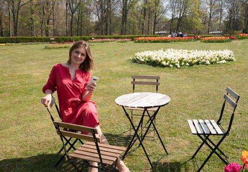 A young beautiful girl in a red dress in a spring park plays with her hair, flirts and takes a selfie on a mobile phone, tulips bloom and people walk in the park, pamper themselves.High quality photo