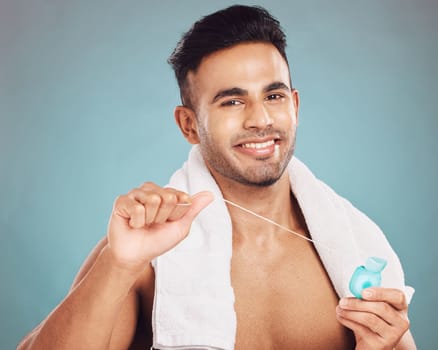 Portrait of one smiling young indian man with towel around neck flossing his teeth after a shower against a blue studio background. Happy guy cleaning his mouth for better oral and dental hygiene. Fl.