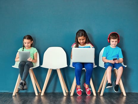 Growing up in a wireless world. Studio shot of kids sitting on chairs and using wireless technology against a blue background