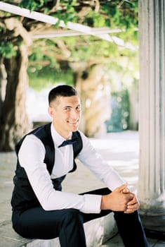 Smiling groom in a black vest sits on the steps near the column in the garden. High quality photo