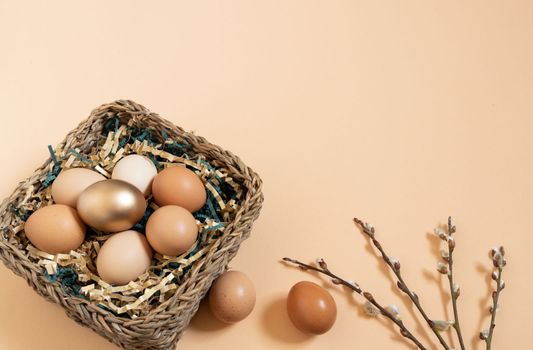 Easter eggs natural and gold in basket with paper filler . Branch of willow catkins. Light pastel orange background with copy space.