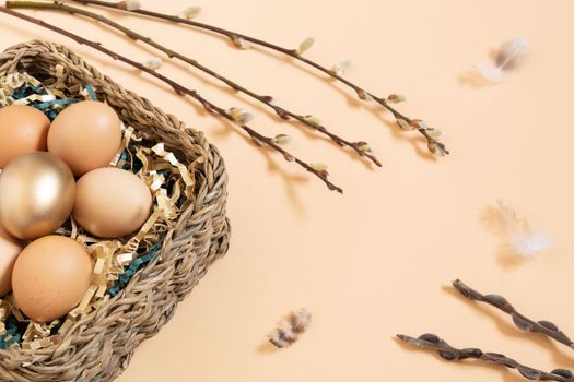 Easter eggs natural and gold in basket with paper filler . Branch of willow catkinsan feather. Light pastel orange background with copy space.